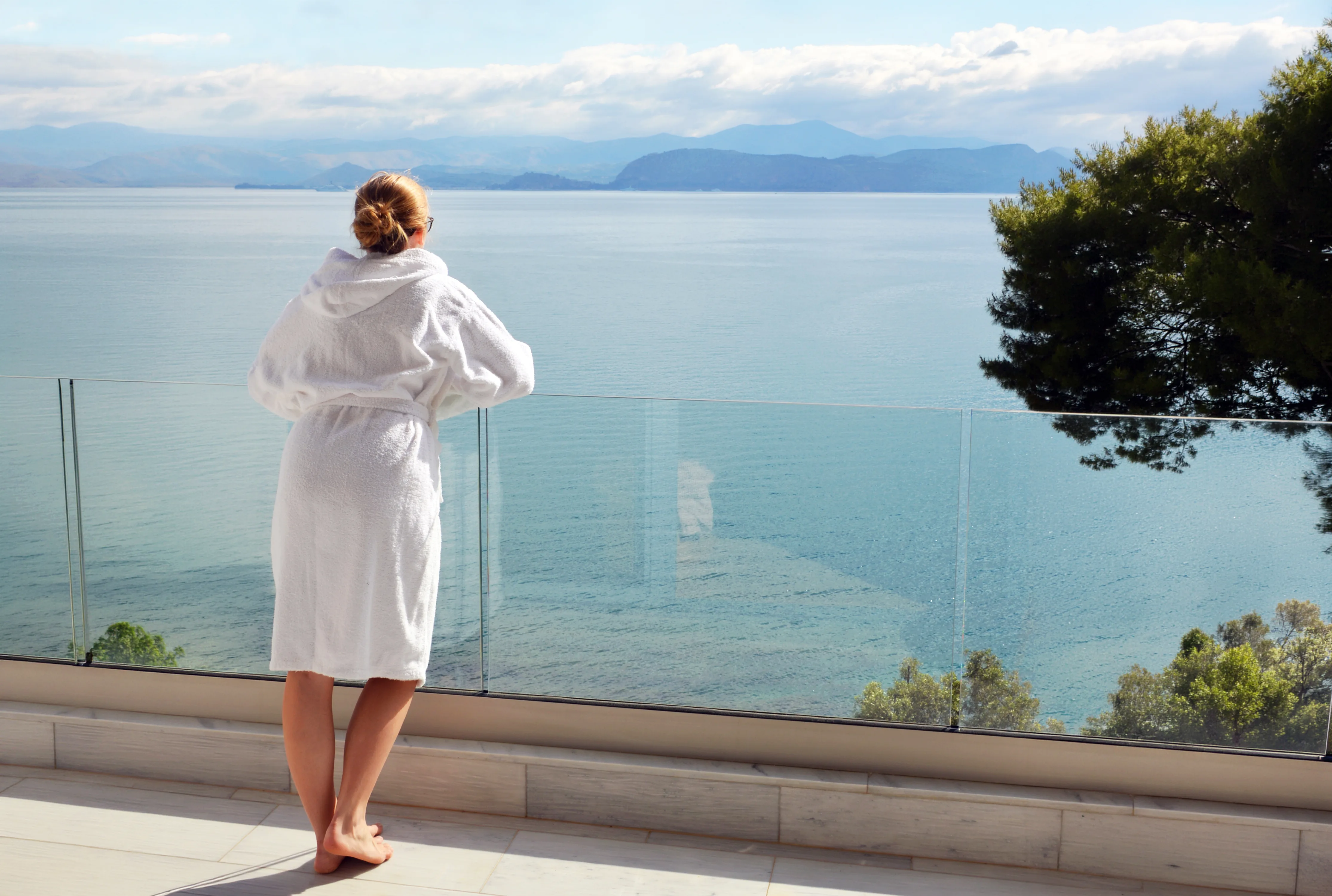 Guest in a bathrobe at a glass balcony rail, looking over the calm gulf and mountains